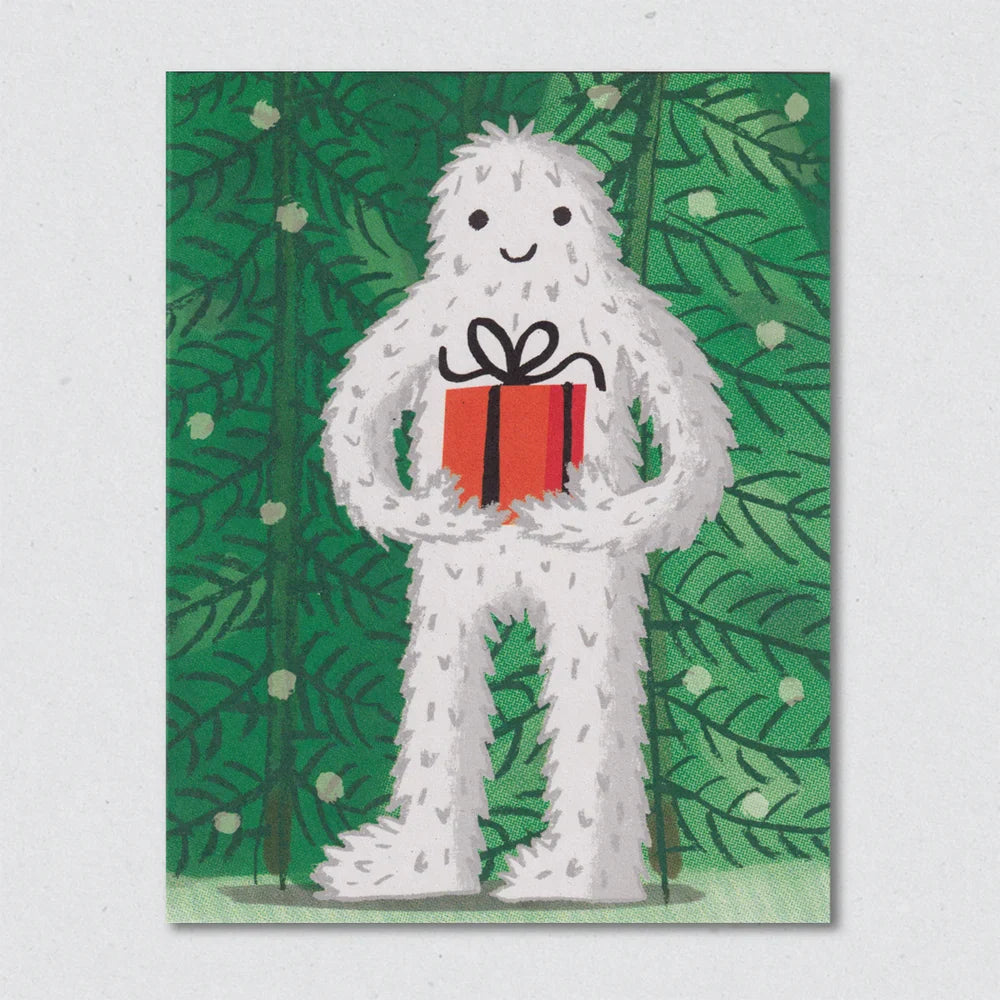 Abominable Snowman Christmas Card by Lisa Jones Studio illustrating the abominable snowman carrying a present in a pine forest in the snow.