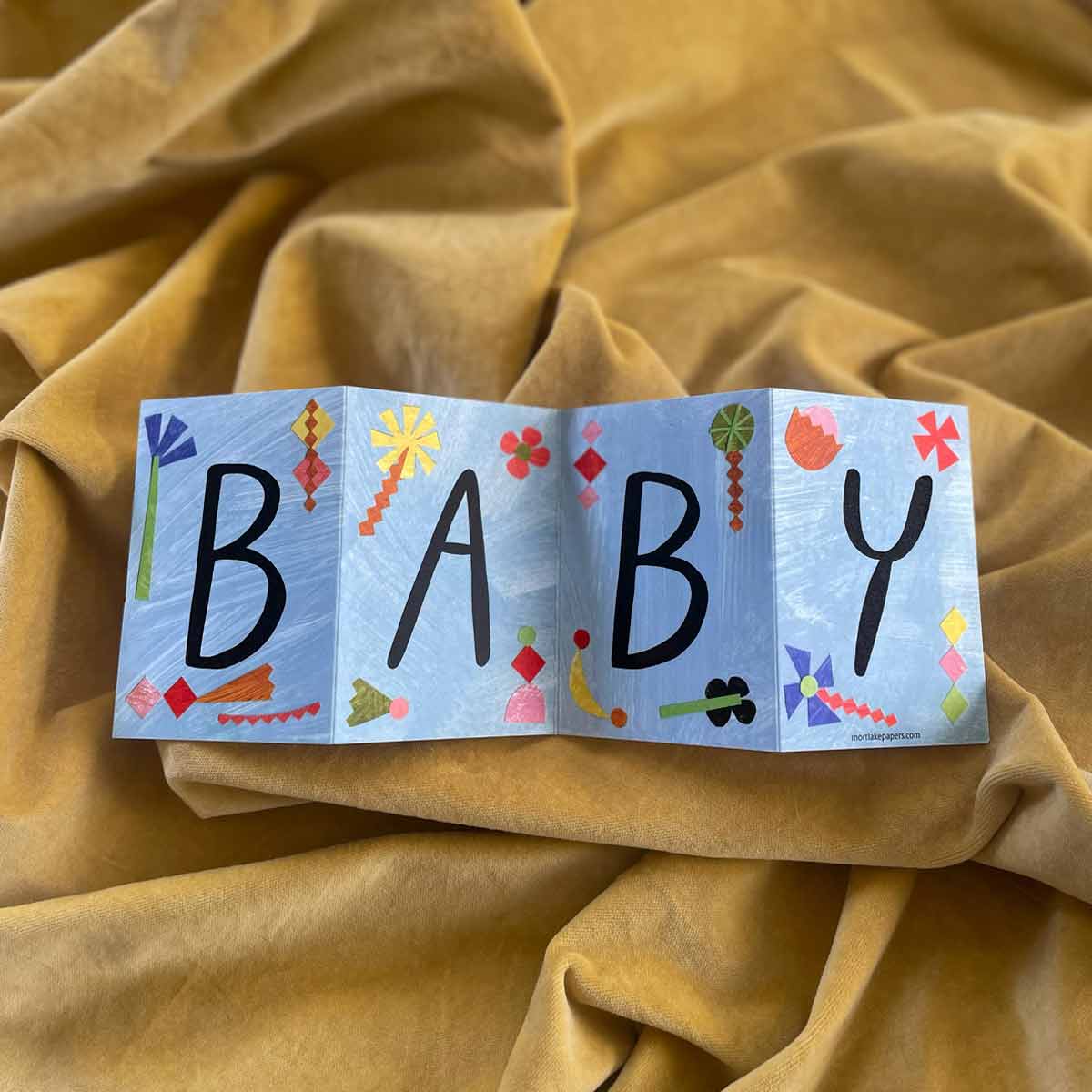 Colorful baby concertina greeting card with 'BABY' on a yellow fabric background by Mortlake Papers by Alice Lickens.