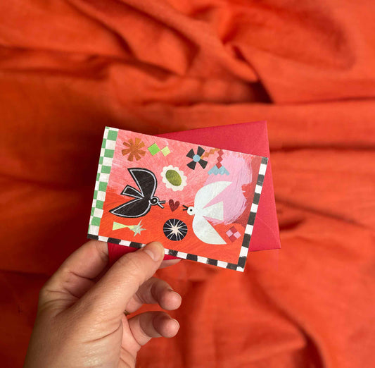 Hand holding a small Birdsong valentines card by Mortlake papers with colorful designs against an orange fabric background.