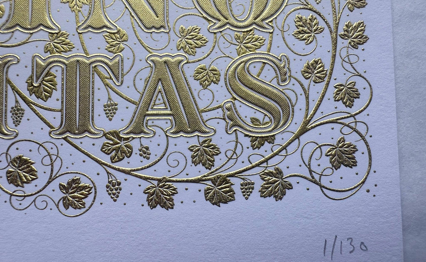 Die stamped print by Seb Lester showing the text In Vino Veritas in fancy lettering and typeface with flourishes and swirls in gold ink on white frost paper. Close up detail.