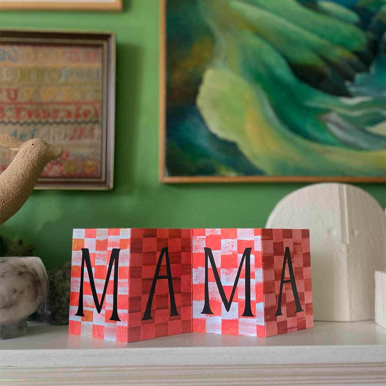 Red checkered concertina greeting card by Mortlake papers with 'MAMA' on a shelf against a green wall with decorative elements.