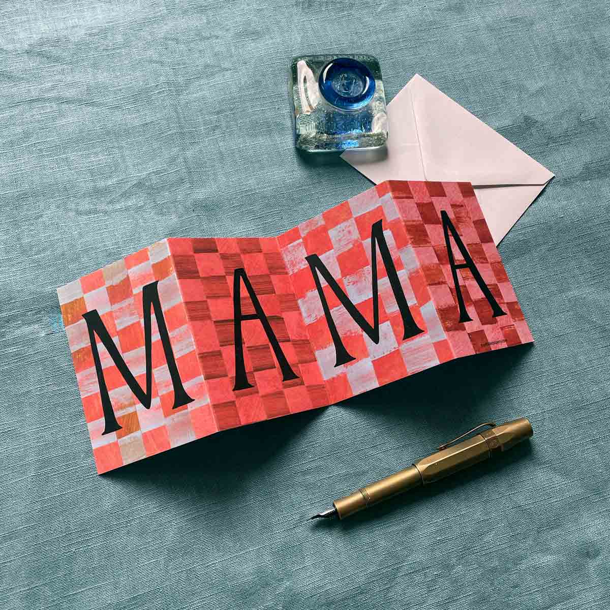 Concertina greeting card by Alice Lickens for Mortlake Papers with 'MAMA' on a checkered pattern, inkwell, envelope, and pen on a textured surface.