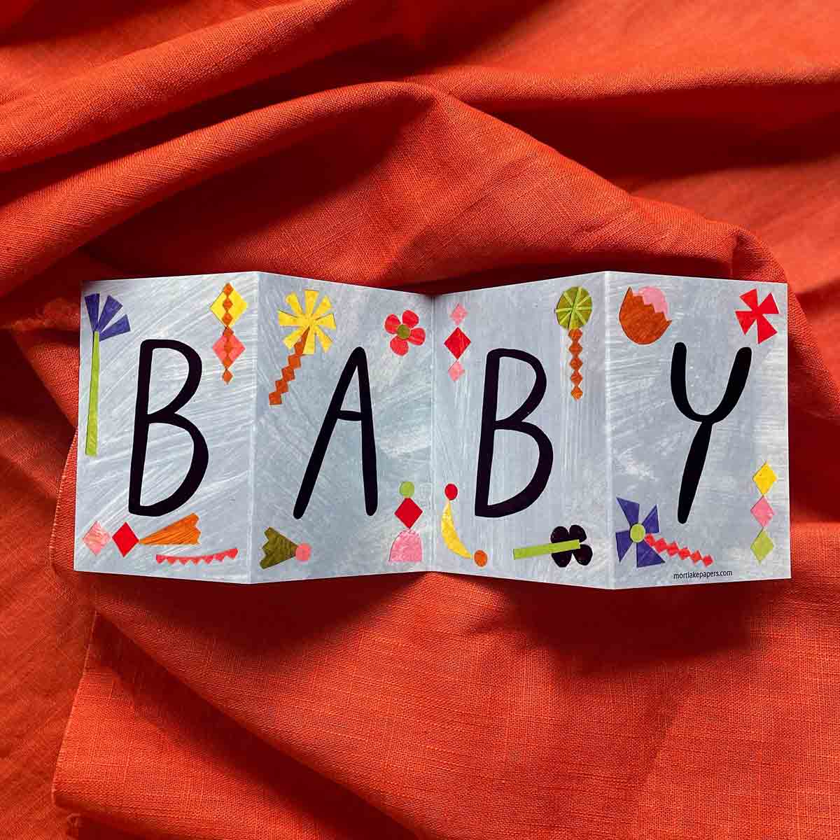 Colorful concertina greeting card by Mortlake Papers by Alice Lickens spelling 'BABY' on an orange fabric background.