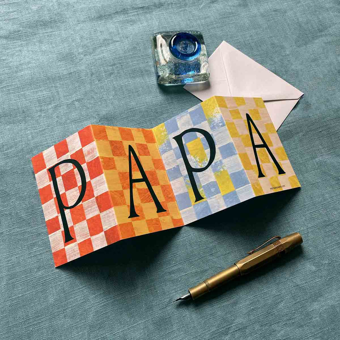 Colorful concertina greeting card by Mortlake Papers with 'PAPA' text on a textured surface with a pen and inkwell.