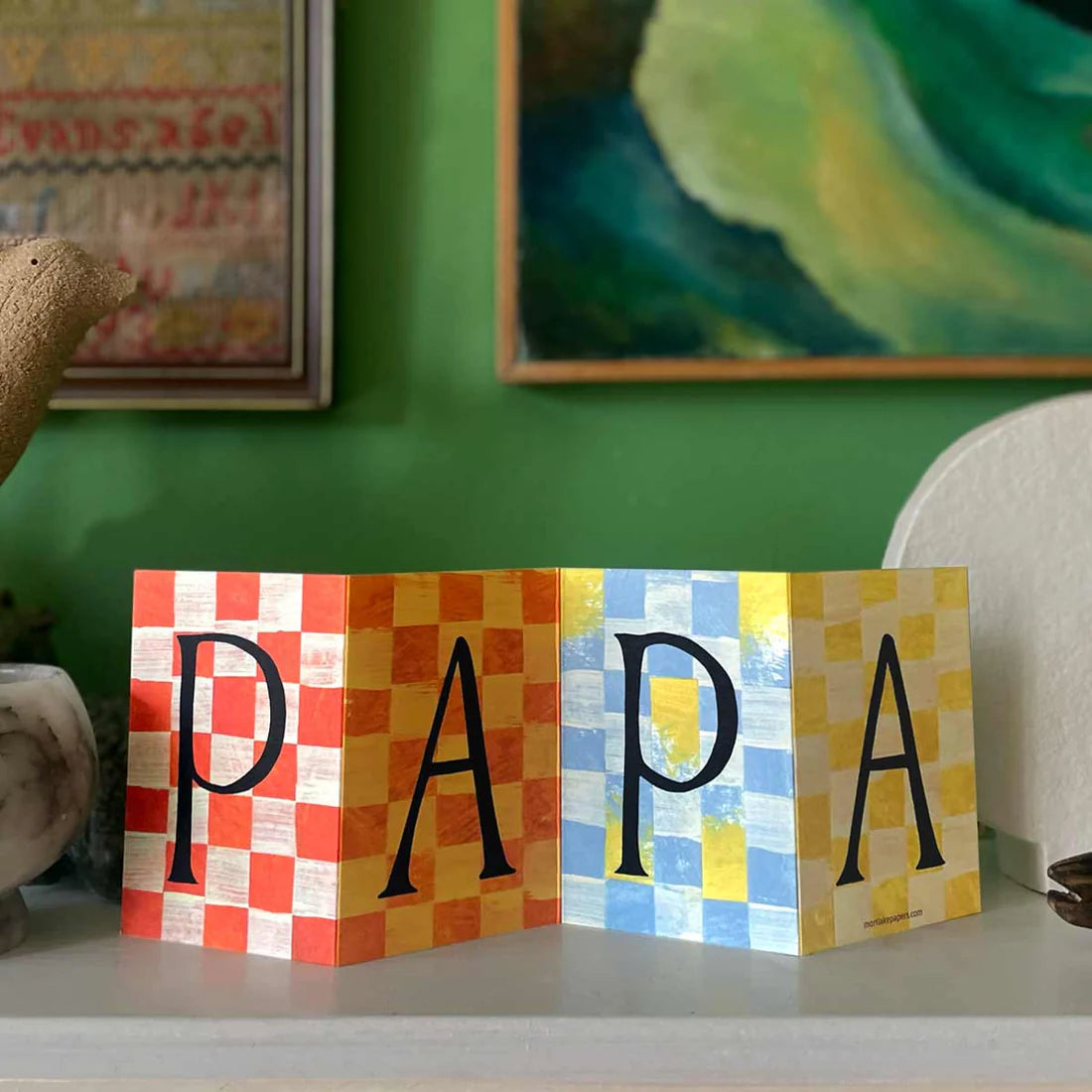 Colorful checkered greeting card by Mortlake Papers spelling 'PAPA' on a shelf with a green wall and framed pictures in the background.
