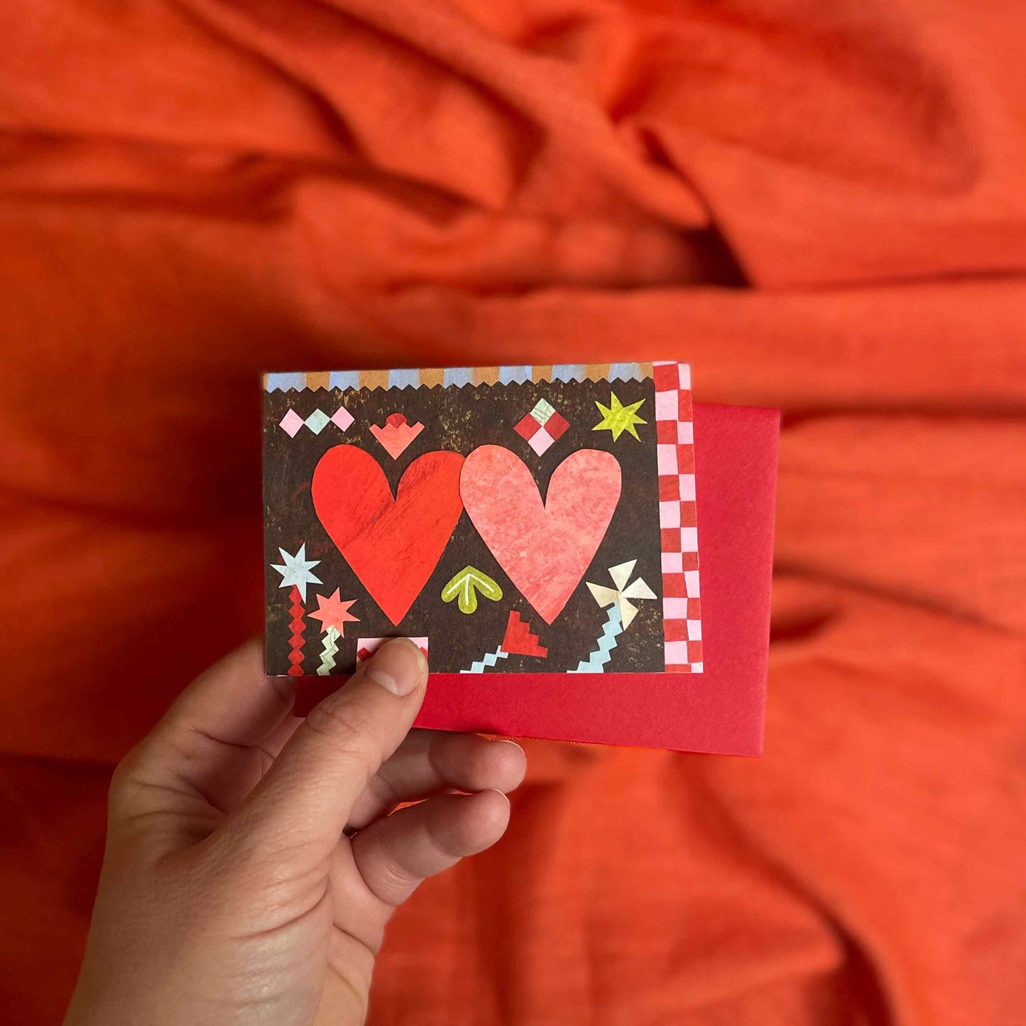 Hand holding a small card, Two Hearts valentines greeting card by Alice Lickens for Mortlake Papers from an original collage, with red hearts against an orange fabric background.