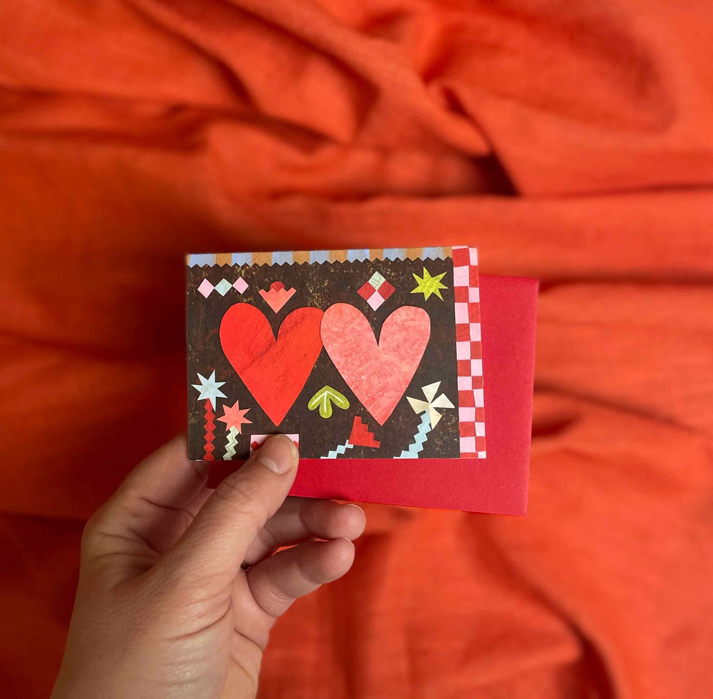 Hand holding a small card, Two Hearts valentines greeting card by Alice Lickens for Mortlake Papers from an original collage, with red hearts against an orange fabric background.