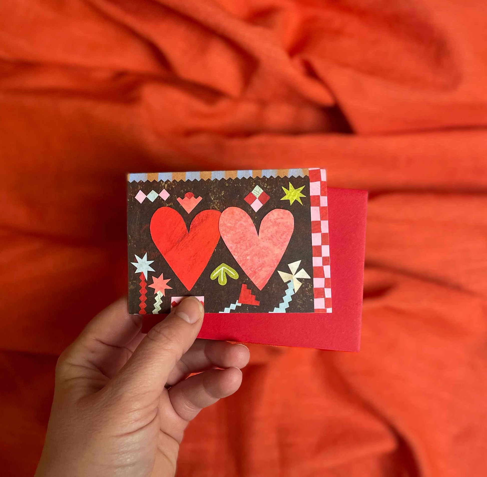 Hand holding a small card, Two Hearts valentines greeting card by Alice Lickens for Mortlake Papers from an original collage, with red hearts against an orange fabric background.