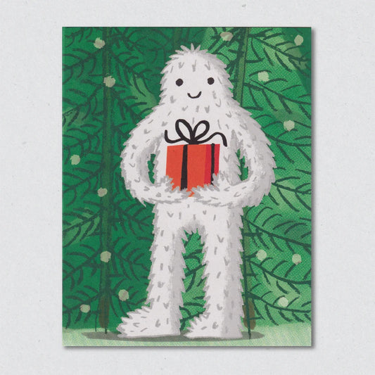 The Abominable Snowman Christmas Card by Lisa Jones Studio illustrating a white creature holding a red gift box against a green leafy background