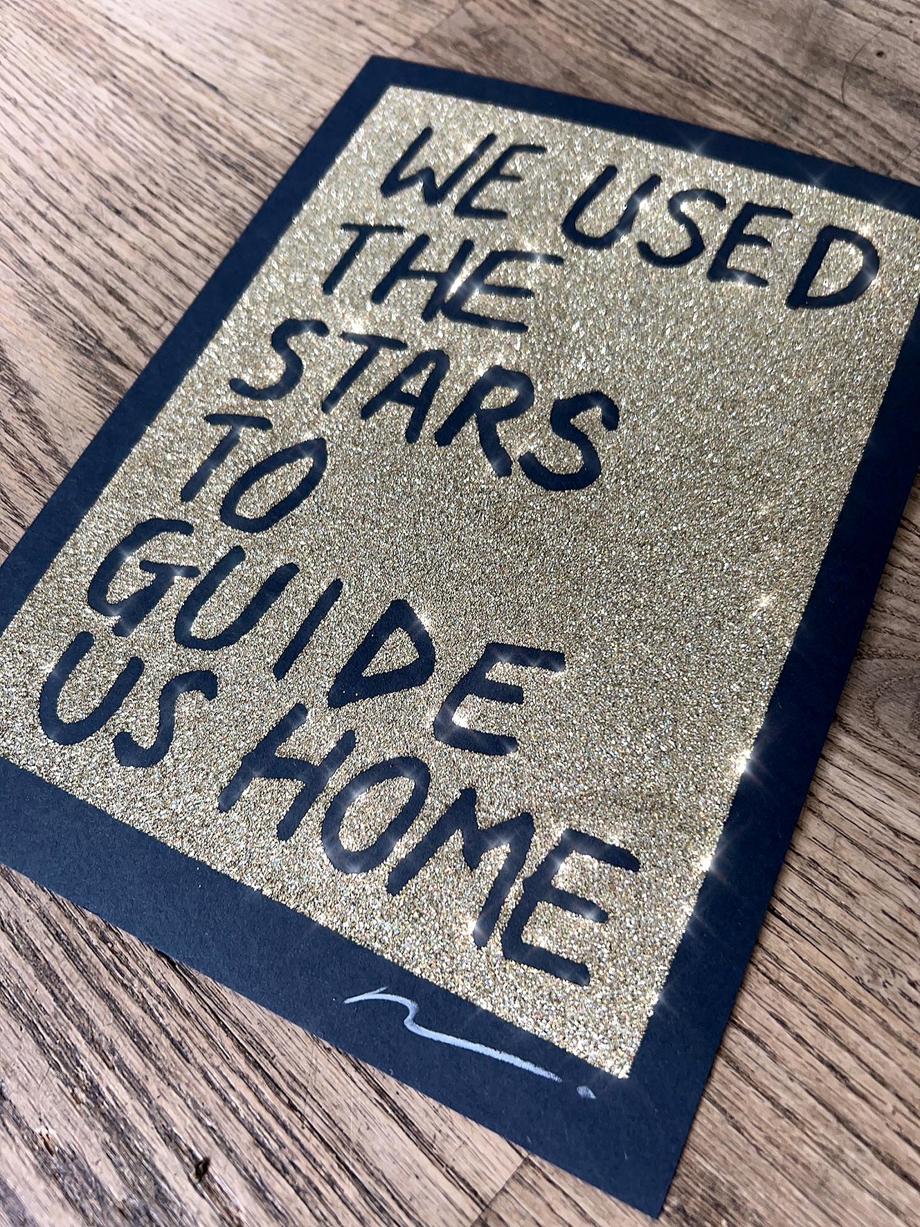 Gold glitter on black paper. Text reads: we used the stars to guide us home in handwritten capitals. By the artist Adam Bridgland and in his signature style