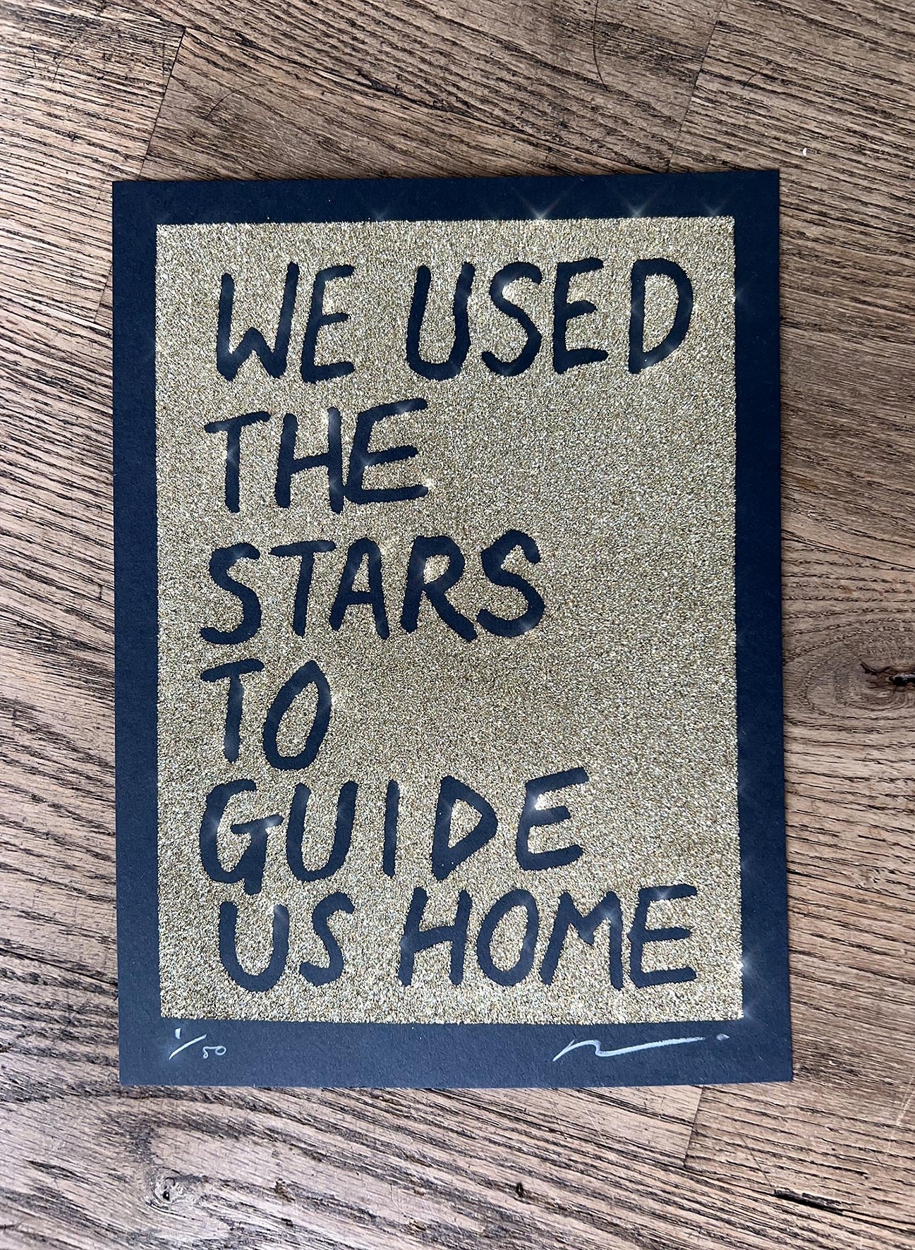 Gold glitter on black paper. Text reads: we used the stars to guide us home in handwritten capitals. By the artist Adam Bridgland and in his signature style