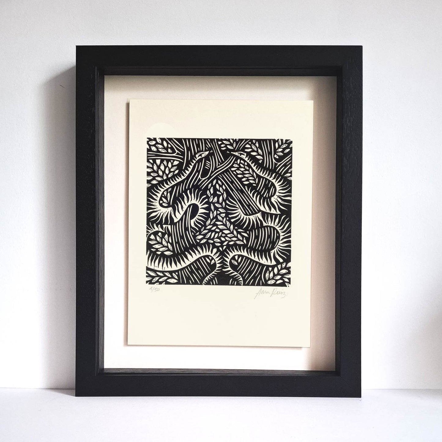 Serpents lino cut print by Anna Ruiz in black ink on off white paper of two snakes curled around tree branches with leaves shown in frame.