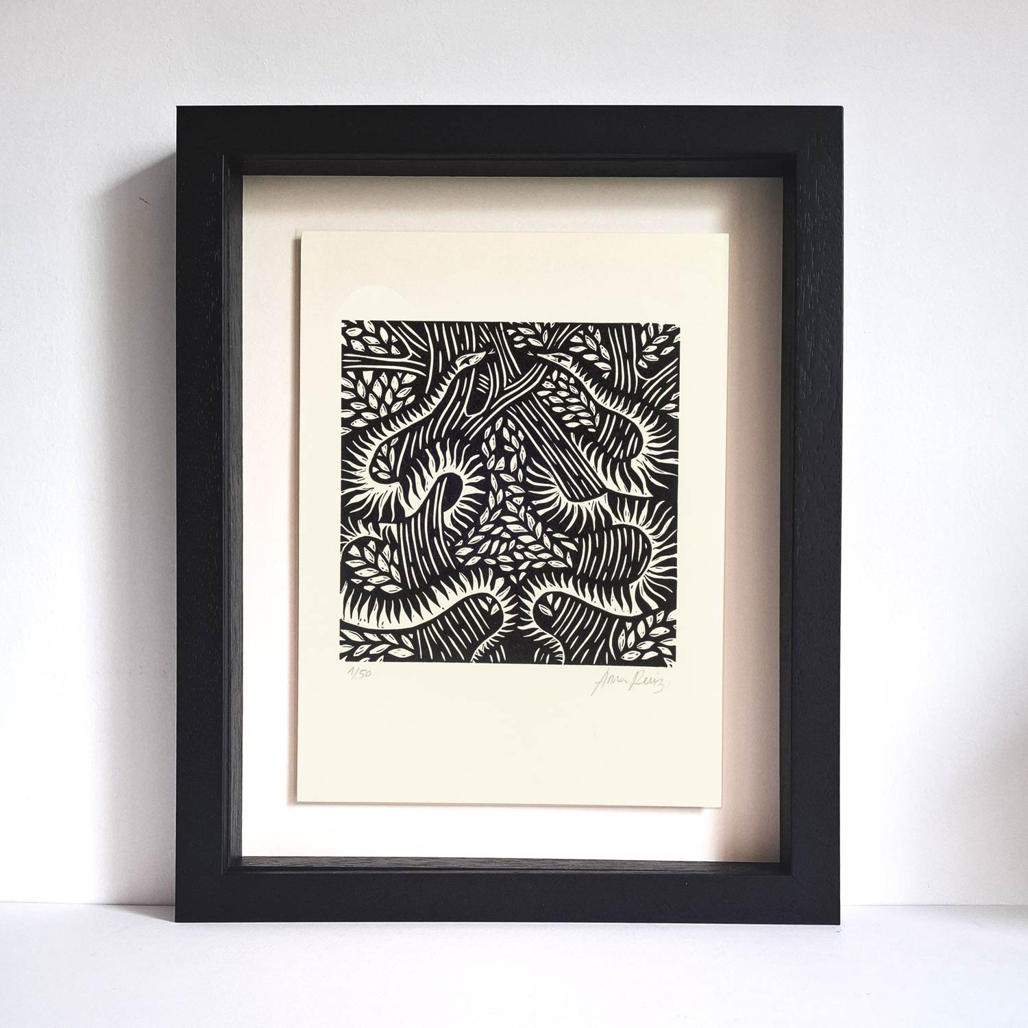 Serpents lino cut print by Anna Ruiz in black ink on off white paper of two snakes curled around tree branches with leaves shown in frame.