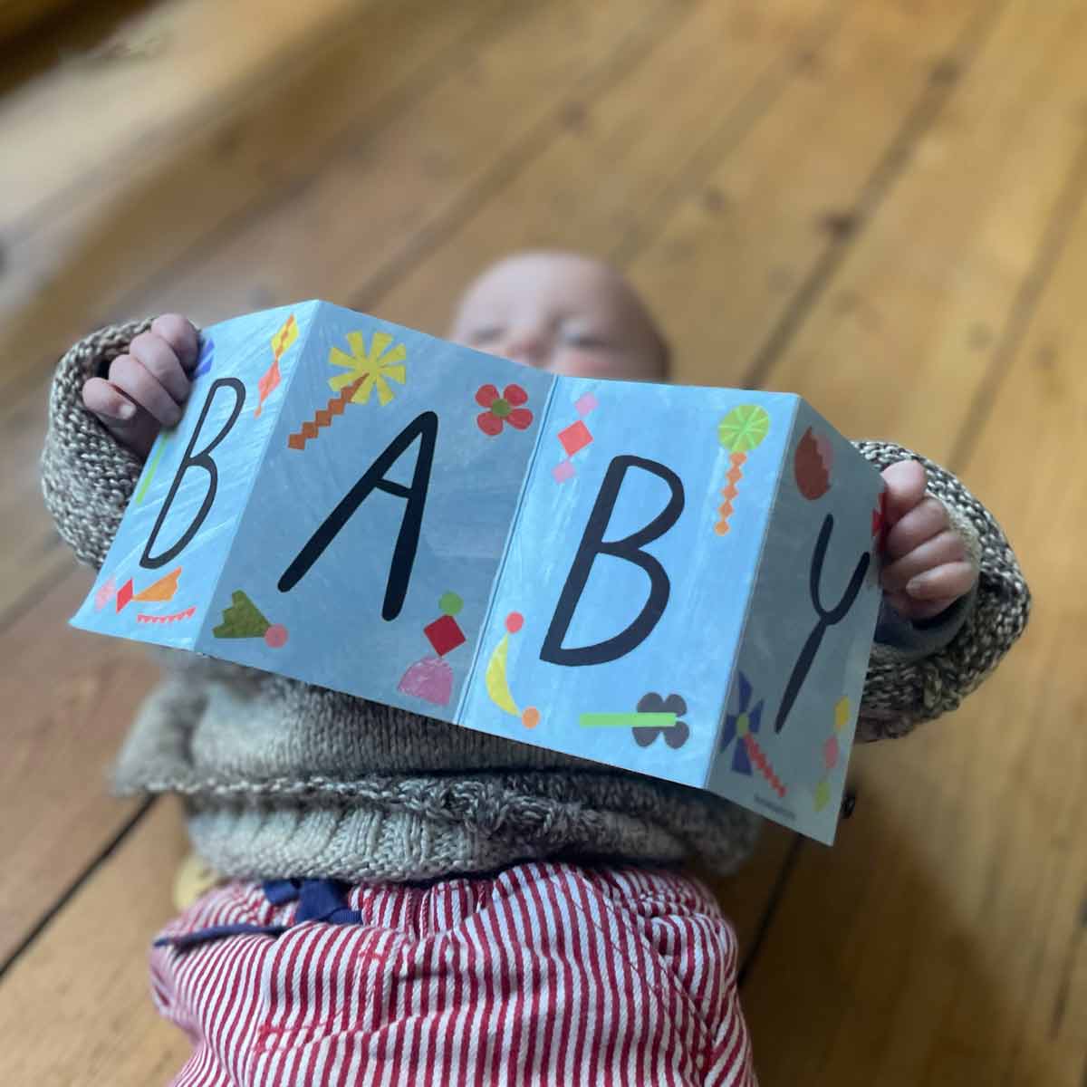 Child holding up greeting card by Mortlake Papers spelling 'BABY' on a wooden floor.