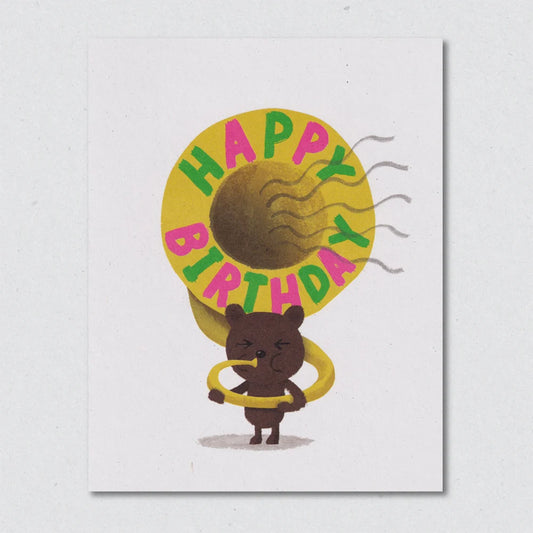Birthday card with illustration of a bear holding a yellow tuba with 'Happy Birthday' text on a white background by Lisa Jones Studio.