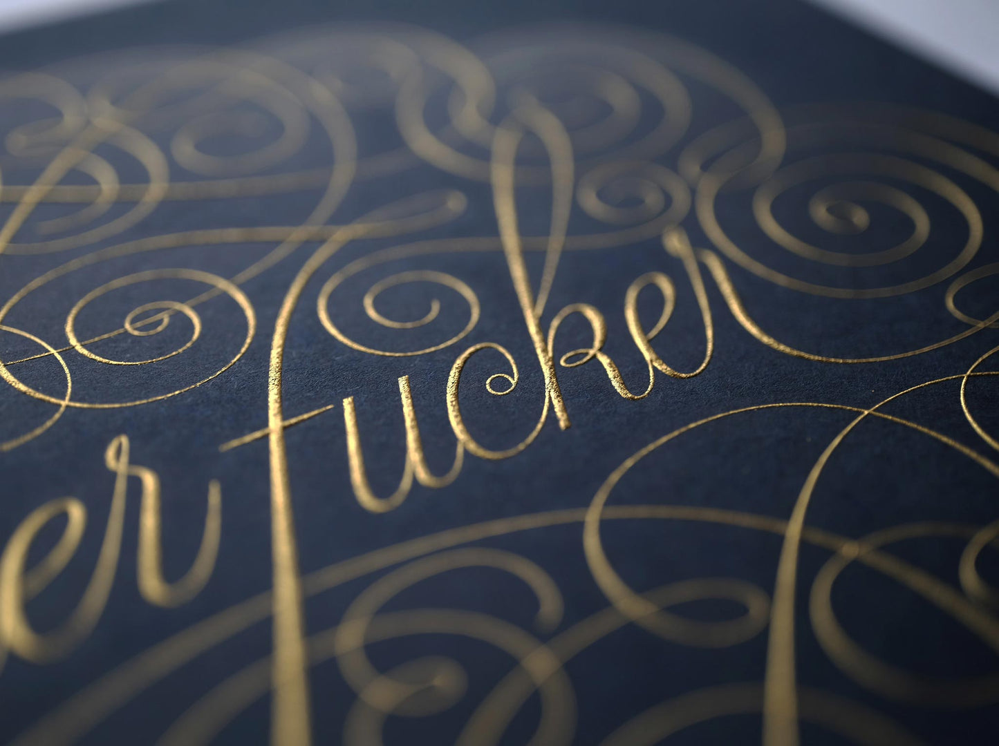 Cursive die stamped artwork by Seb lester. Featuring ornate swirls and swooshes around a swearword. Gold ink on blue paper.