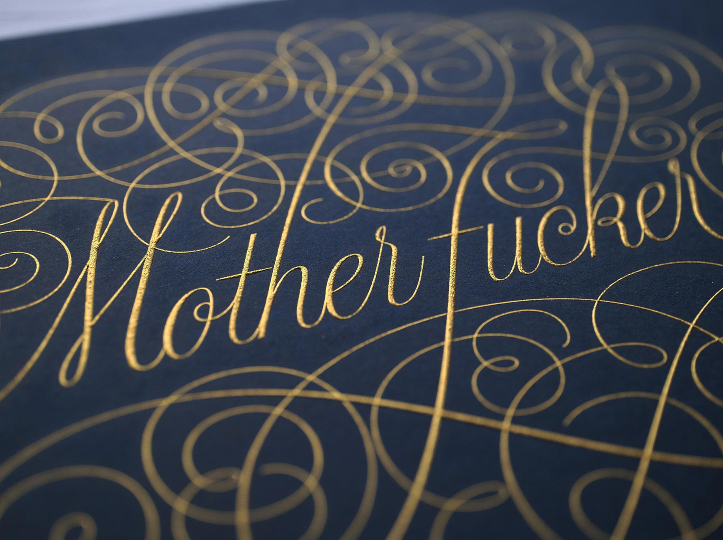 Cursive die stamped artwork by Seb lester. Featuring ornate swirls and swooshes around a swearword. Gold ink on blue paper.