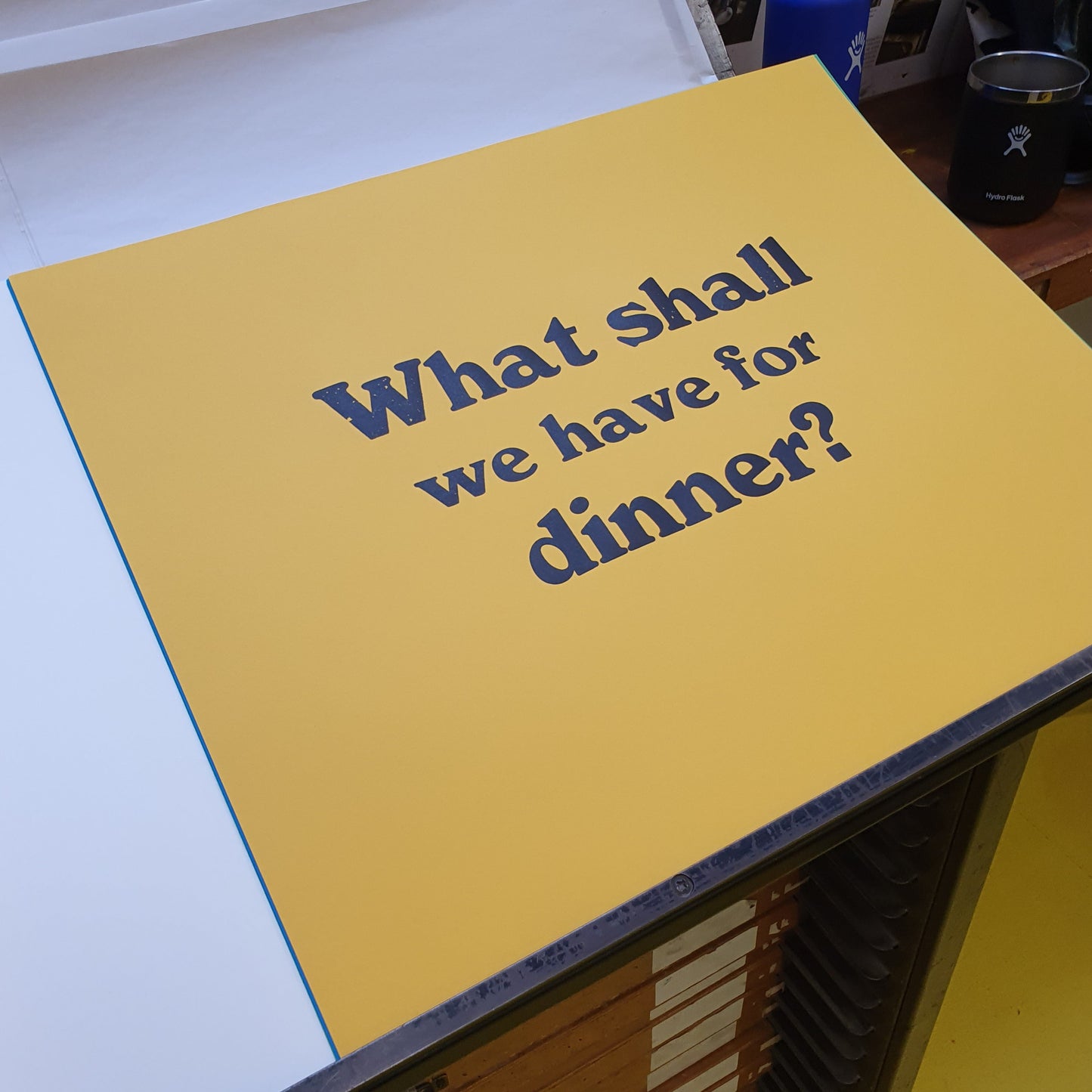 A yellow colored poster with the text 'WHAT SHALL WE HAVE FOR DINNER?' printed in black. The poster is placed on a table with a blurred background.