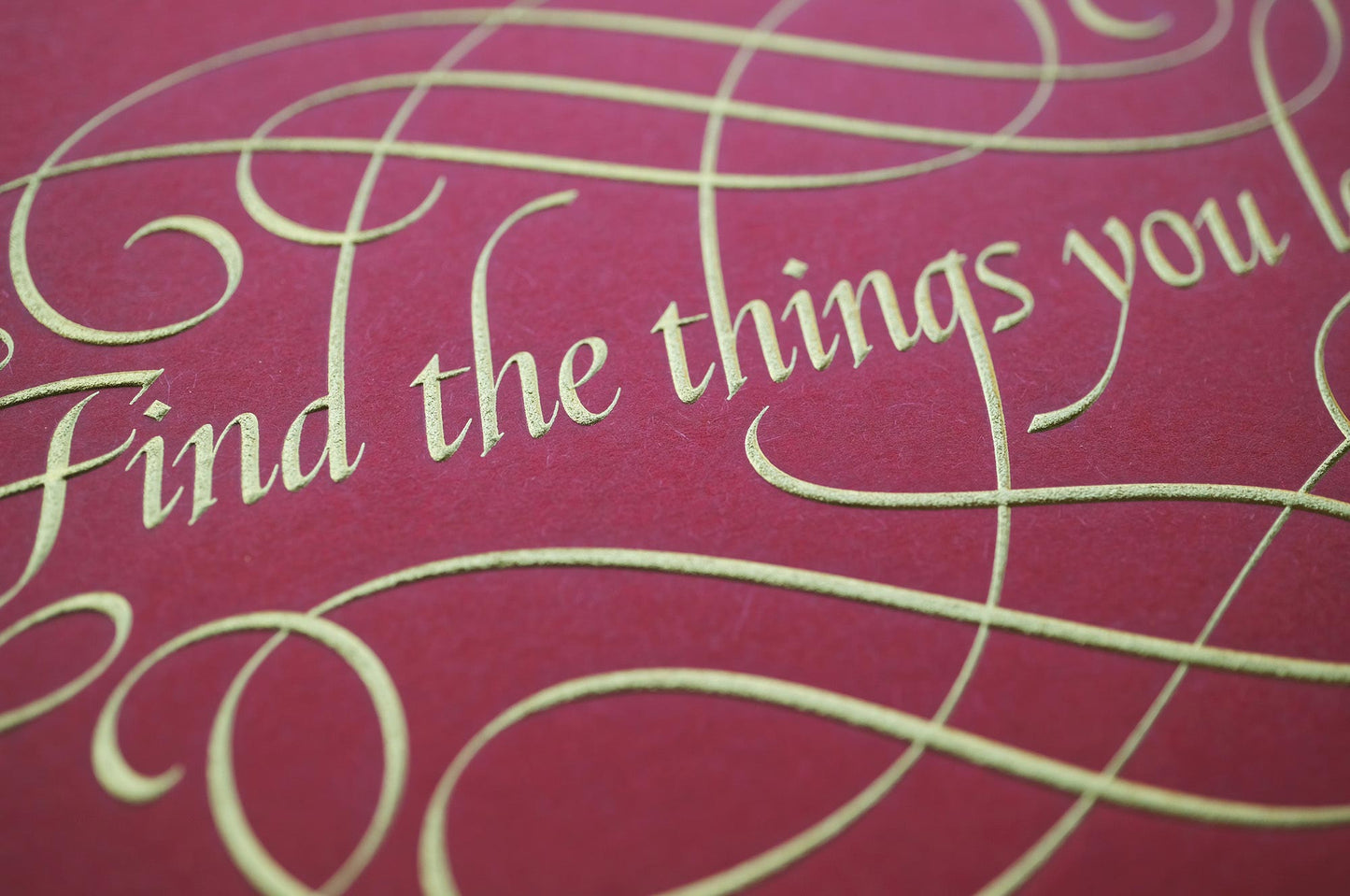 Find the things print by Seb Lester. Die stamp with raised impression. Illustrates the text Find the things you love with flourishes and swirls in gold on red. Close up detail.