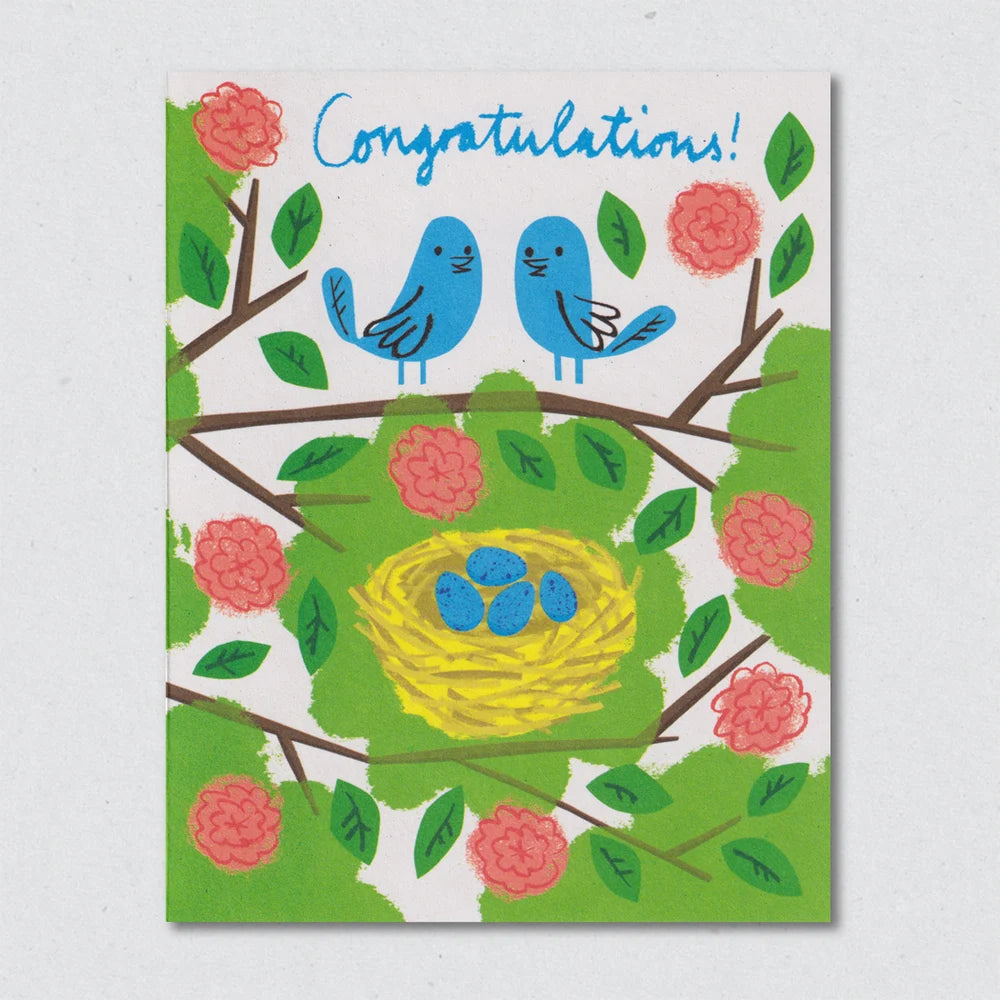 Greeting card with blue birds, a yellow nest, and pink flowers on a white background by Lisa Jones Studio.