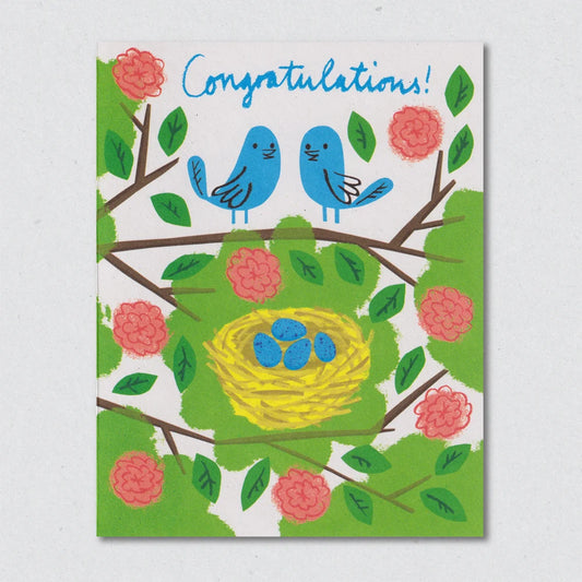 Greeting card with blue birds, a yellow nest, and pink flowers on a white background by Lisa Jones Studio.
