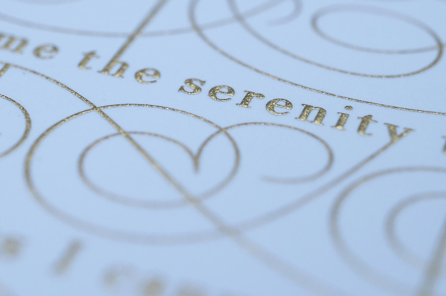 The serenity prayer die stamp print by Seb Lester in gold on white. Features text: "god grant me the serenity to accept the things I cannot change, courage to change the things I can and wisdom to know the different. Type in ornate font and flourishes and swirls around the text. Close up detail.