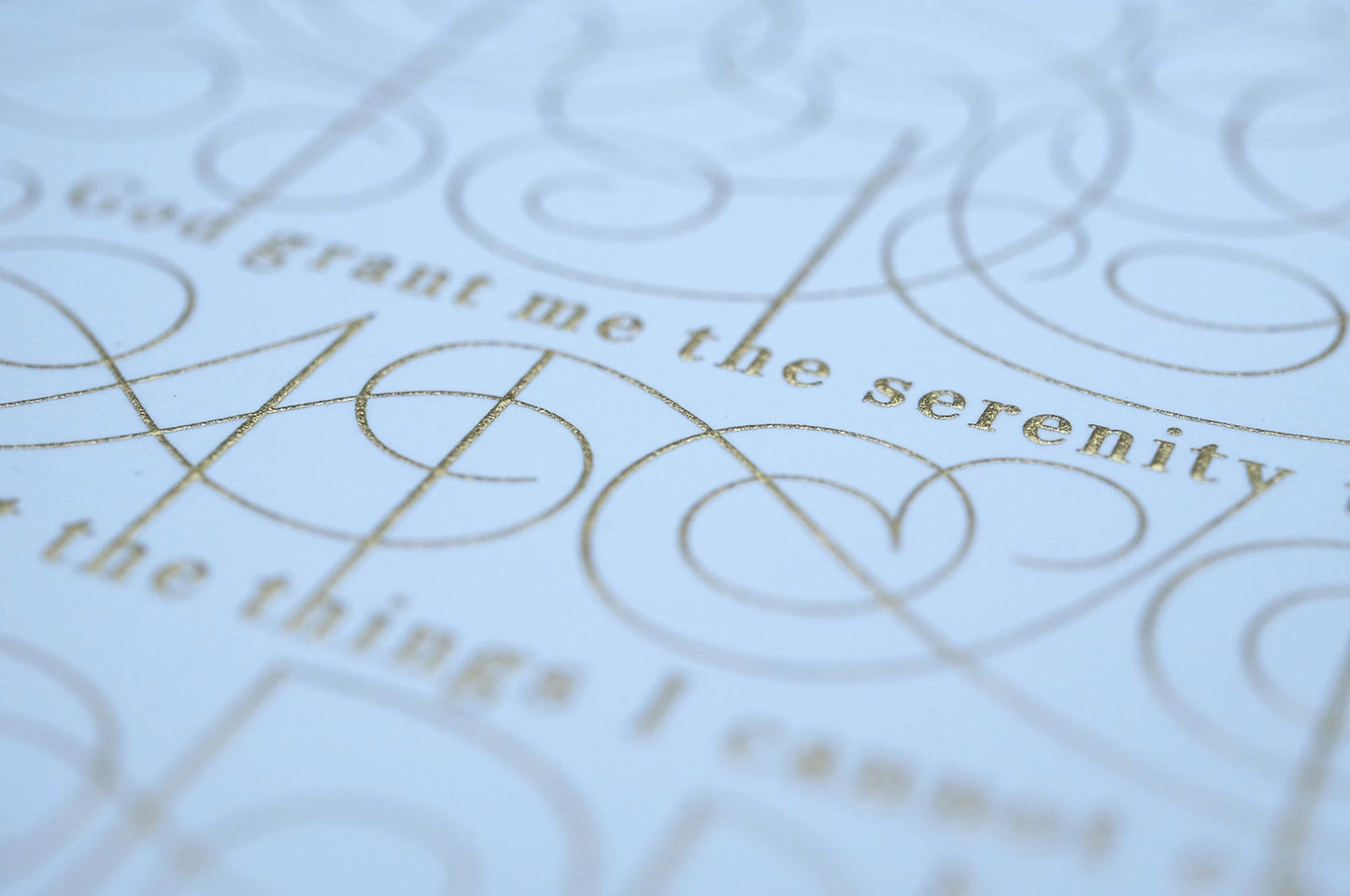 The serenity prayer die stamp print by Seb Lester in gold on white. Features text: "god grant me the serenity to accept the things I cannot change, courage to change the things I can and wisdom to know the different. Type in ornate font and flourishes and swirls around the text. Close up detail.