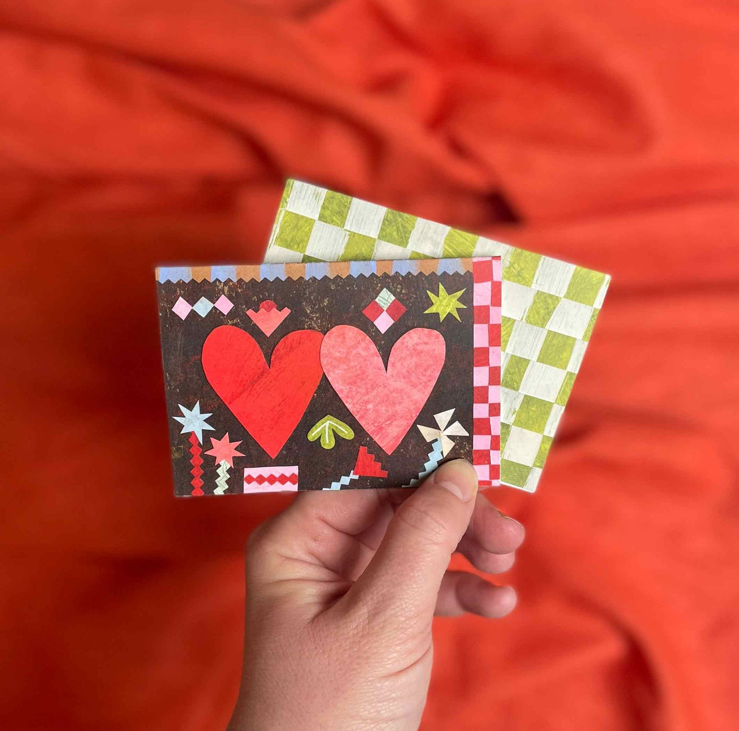 Hand holding two small cards with heart designs against an orange fabric background by Alice Lickens for Mortlake Papers.
