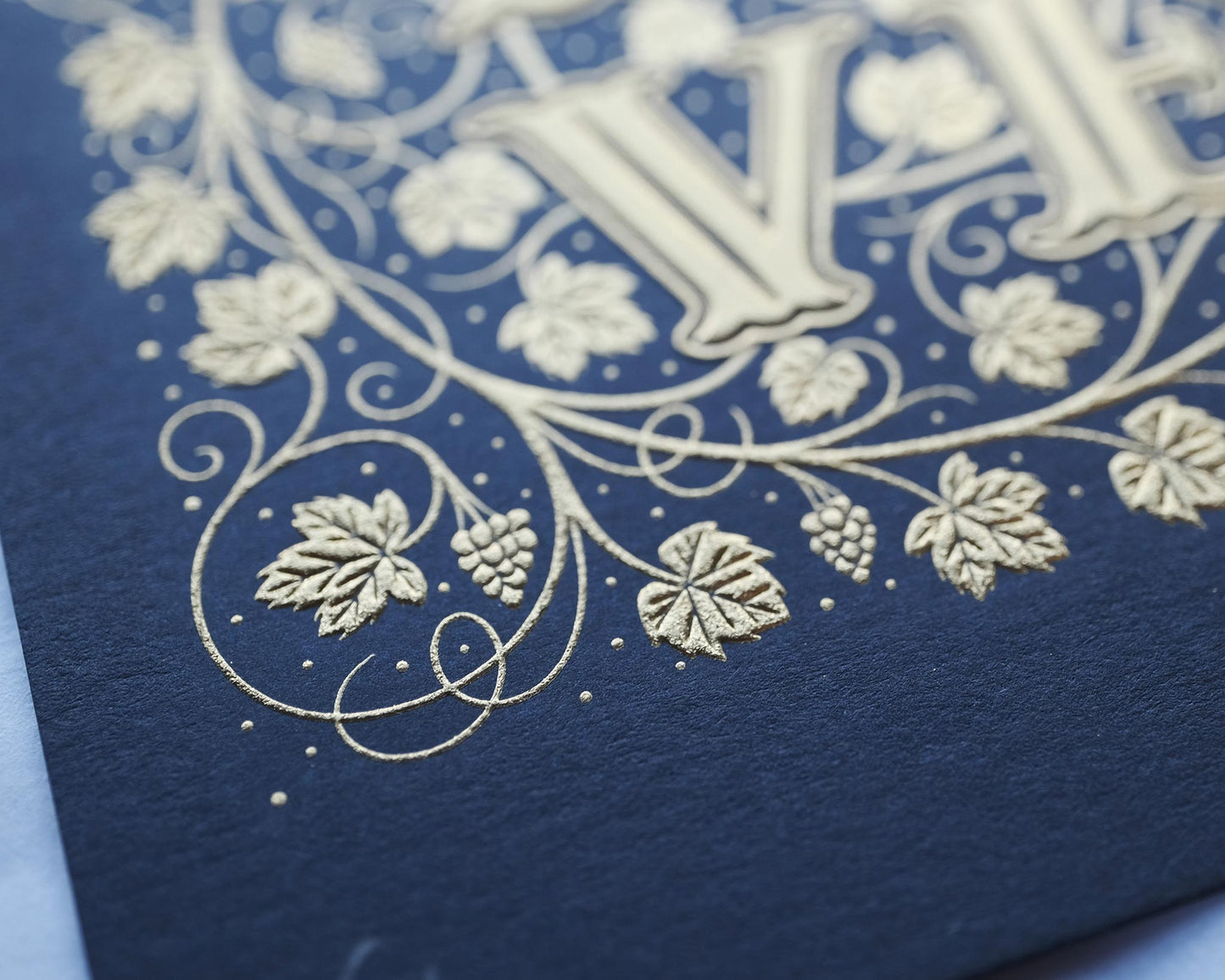 Die stamped print by Seb Lester showing the text In Vino Veritas in fancy lettering and typeface with flourishes and swirls in gold ink on blue paper. Close up detail.