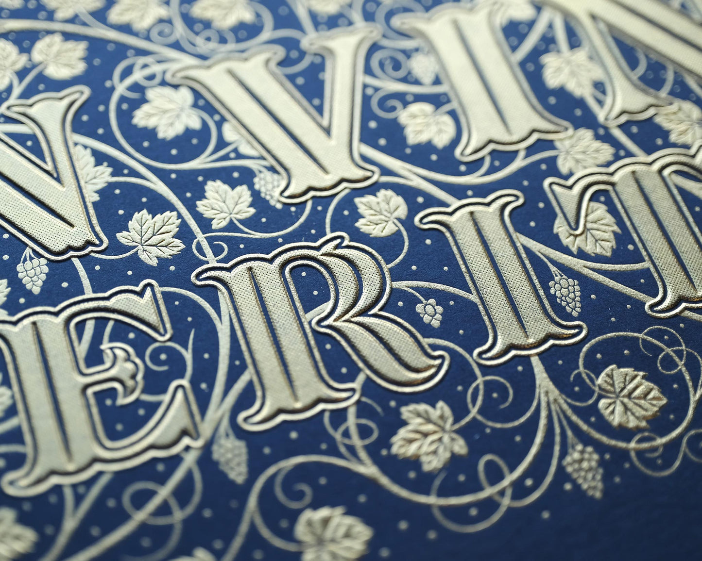 Die stamped print by Seb Lester showing the text In Vino Veritas in fancy lettering and typeface with flourishes and swirls in gold ink on blue paper. Close up detail.