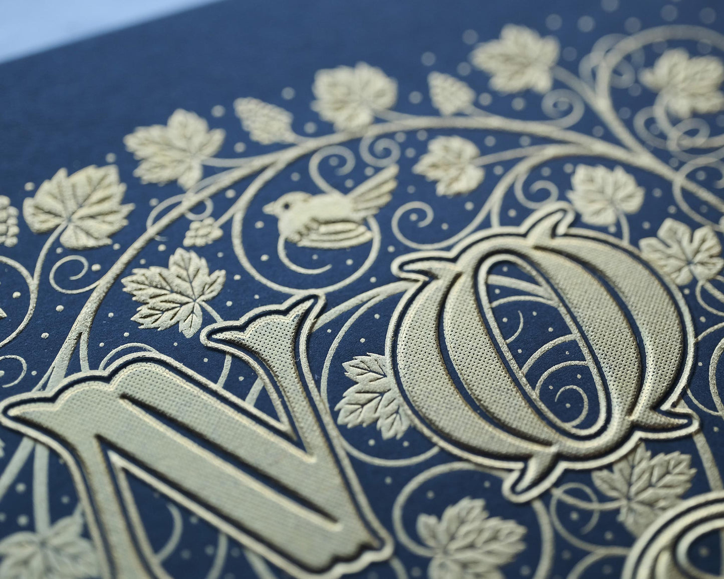 Die stamped print by Seb Lester showing the text In Vino Veritas in fancy lettering and typeface with flourishes and swirls in gold ink on blue paper. Close up detail.