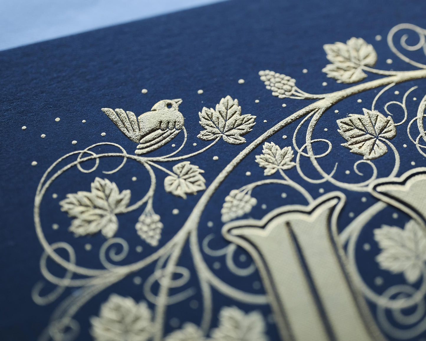 Die stamped print by Seb Lester showing the text In Vino Veritas in fancy lettering and typeface with flourishes and swirls in gold ink on blue paper. Close up detail.