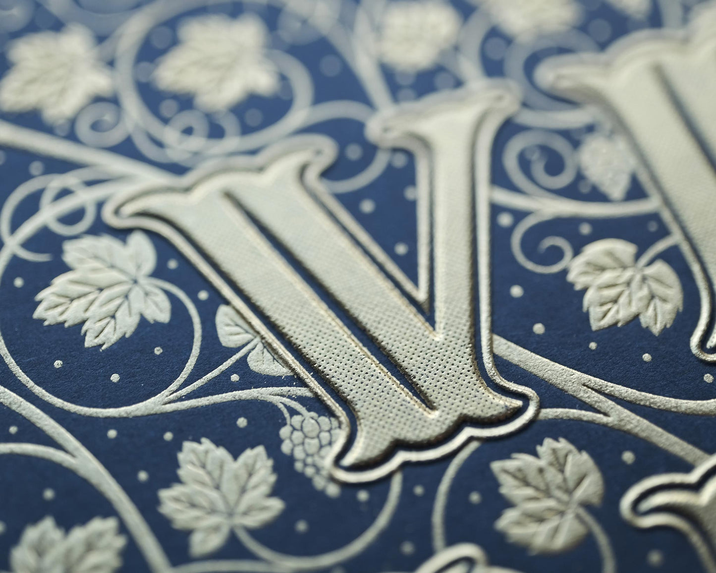 Die stamped print by Seb Lester showing the text In Vino Veritas in fancy lettering and typeface with flourishes and swirls in gold ink on blue paper. Close up detail.