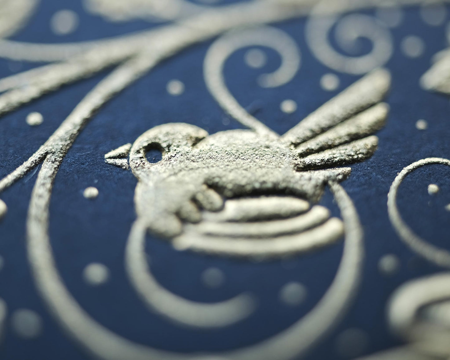 Die stamped print by Seb Lester showing the text In Vino Veritas in fancy lettering and typeface with flourishes and swirls in gold ink on blue paper. Close up detail.