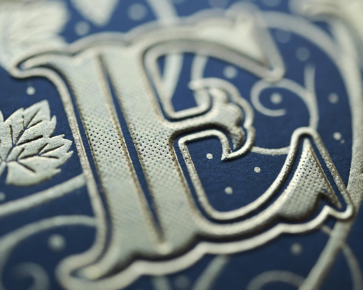 Die stamped print by Seb Lester showing the text In Vino Veritas in fancy lettering and typeface with flourishes and swirls in gold ink on blue paper. Close up detail.