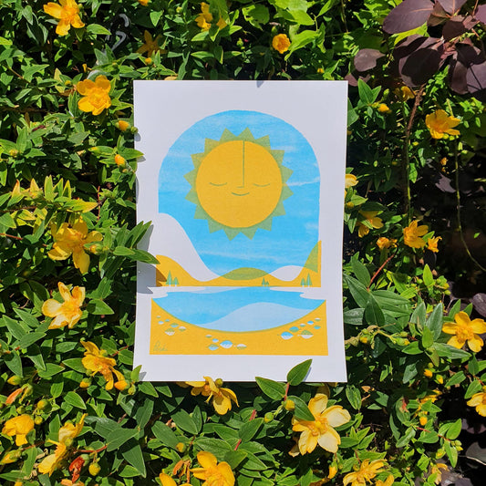 A risograph print titled 'Hello Sunshine' placed on a bed of yellow flowers, depicting a sun over a blue lake, held by someone.