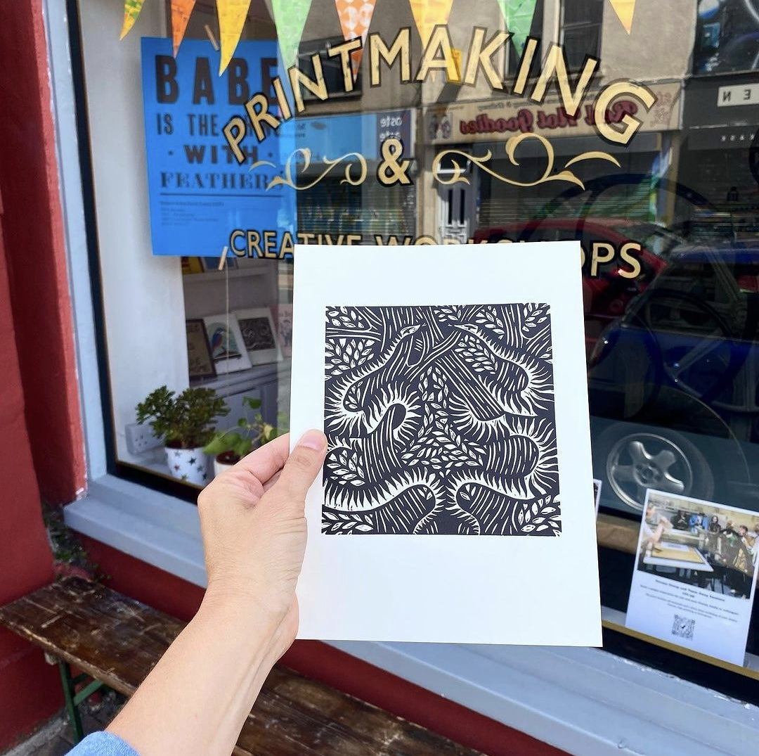 Serpents lino cut print by Anna Ruiz in black ink on off white paper of two snakes curled around tree branches with leaves in the window of Frankenstein Press 