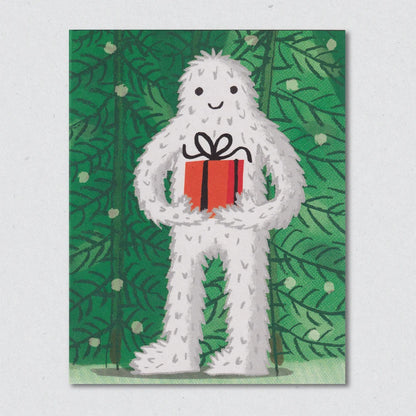The Abominable Snowman Christmas Card by Lisa Jones Studio illustrating a white creature holding a red gift box against a green leafy background