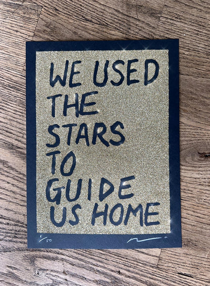 Gold glitter on black paper. Text reads: we used the stars to guide us home in handwritten capitals. By the artist Adam Bridgland and in his signature style
