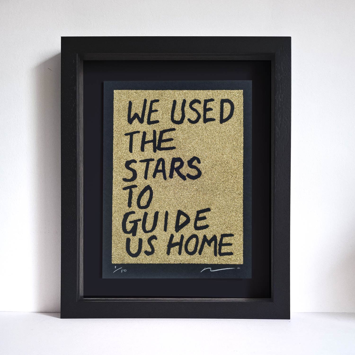 Gold glitter on black paper framed original art screen print. Text reads: we used the stars to guide us home in handwritten capitals. By the artist Adam Bridgland and in his signature style