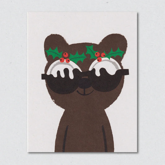 Pudding Face Christmas card by Lisa Jones Studio featuring an illustration of a brown bear wearing sunglasses with holly leaves and berries on a white background.