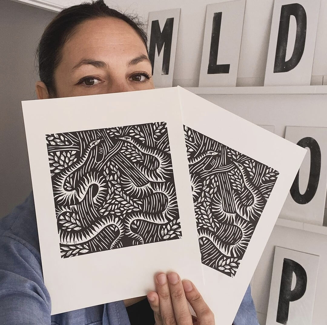 Serpents lino cut print by Anna Ruiz in black ink on off white paper of two snakes curled around tree branches with leaves being held by Anna