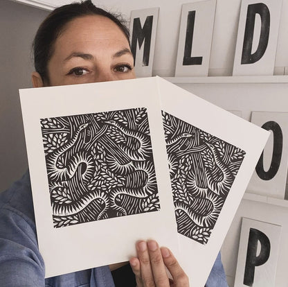 Serpents lino cut print by Anna Ruiz in black ink on off white paper of two snakes curled around tree branches with leaves being held by Anna