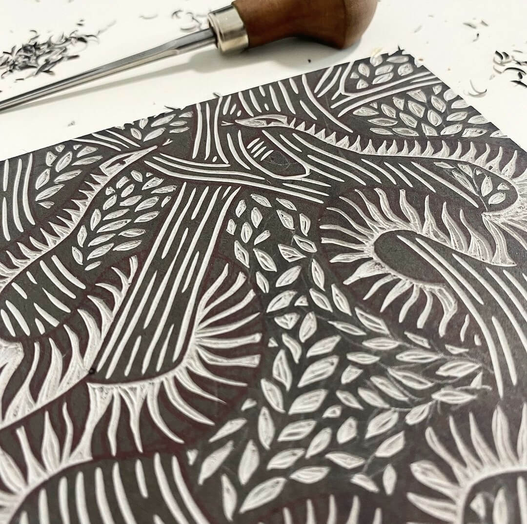 Serpents lino cut print by Anna Ruiz in black ink on off white paper of two snakes curled around tree branches with leaves.