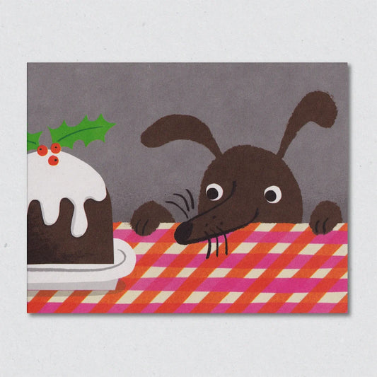 Single Serving Christmas Card by Lisa Jones Studio illustrating a dog that's just about to devour a Christmas pudding, that is sat on a gingham tablecloth, in one bite. The dogs head and paws are rising up from the table.