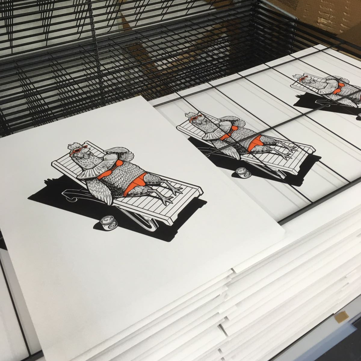 Sunbathing Chicken screen prints by Matthew Green on the drying rack featuring an illustration of a chicken on a sun lounger in black and white wearing an orange bikini.