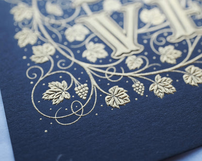 Die stamped print by Seb Lester showing the text In Vino Veritas in fancy lettering and typeface with flourishes and swirls in gold ink on blue paper. Close up detail.